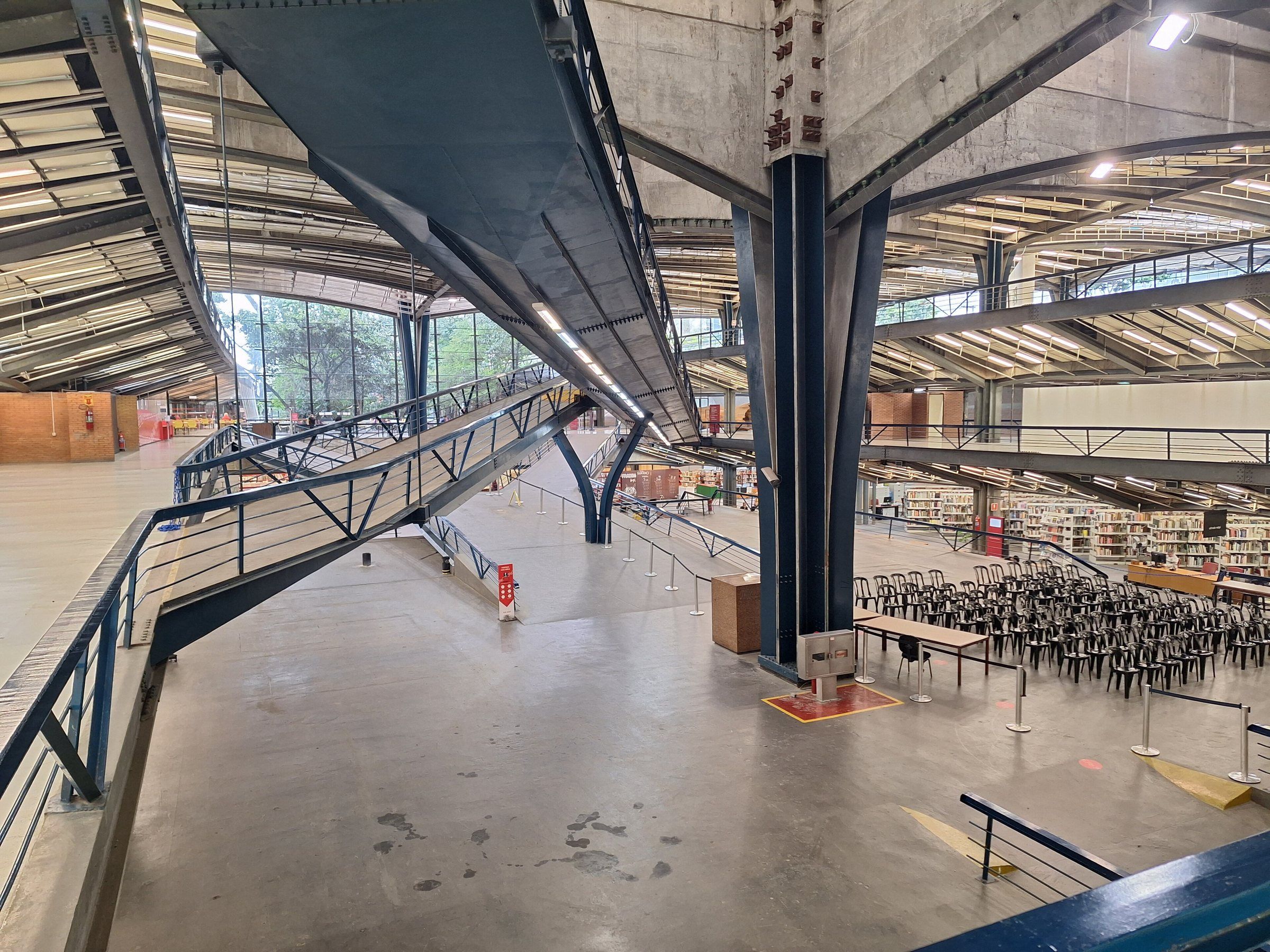 Interior amplo com estrutura metálica azul, rampas que descem entre pavimentos, estantes de biblioteca e fileiras de cadeiras.