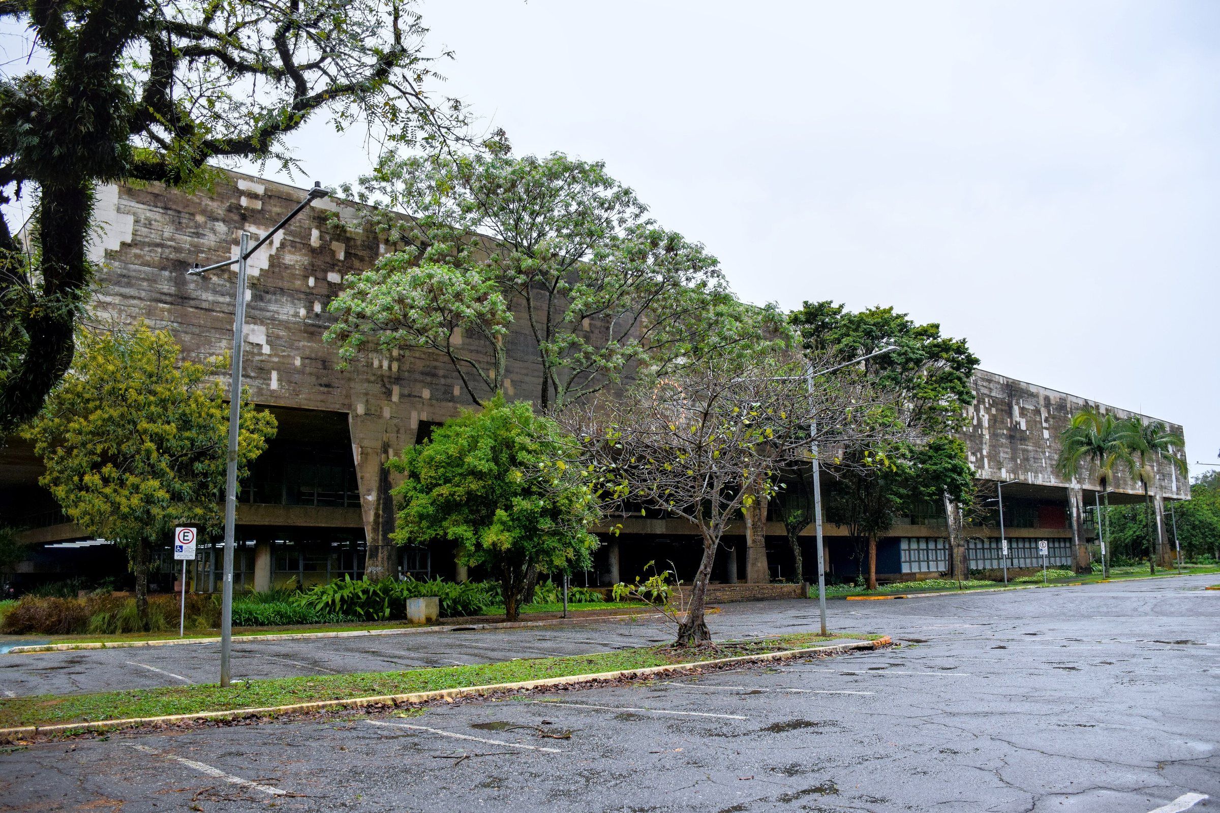 Longa fachada de concreto manchado pelo tempo, parcialmente escondida por árvores, diante de um estacionamento vazio em dia nublado.