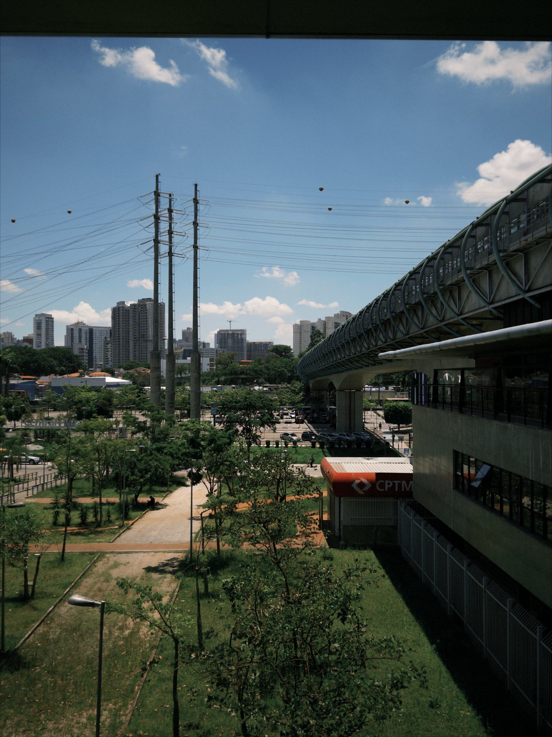 Vista do alto, sob a borda de uma laje de concreto: torres de transmissão e prédios contra o céu azul, uma via elevada em curva à direita, jardins com caminhos de terra e, entre eles, uma cobertura vermelha da CPTM.