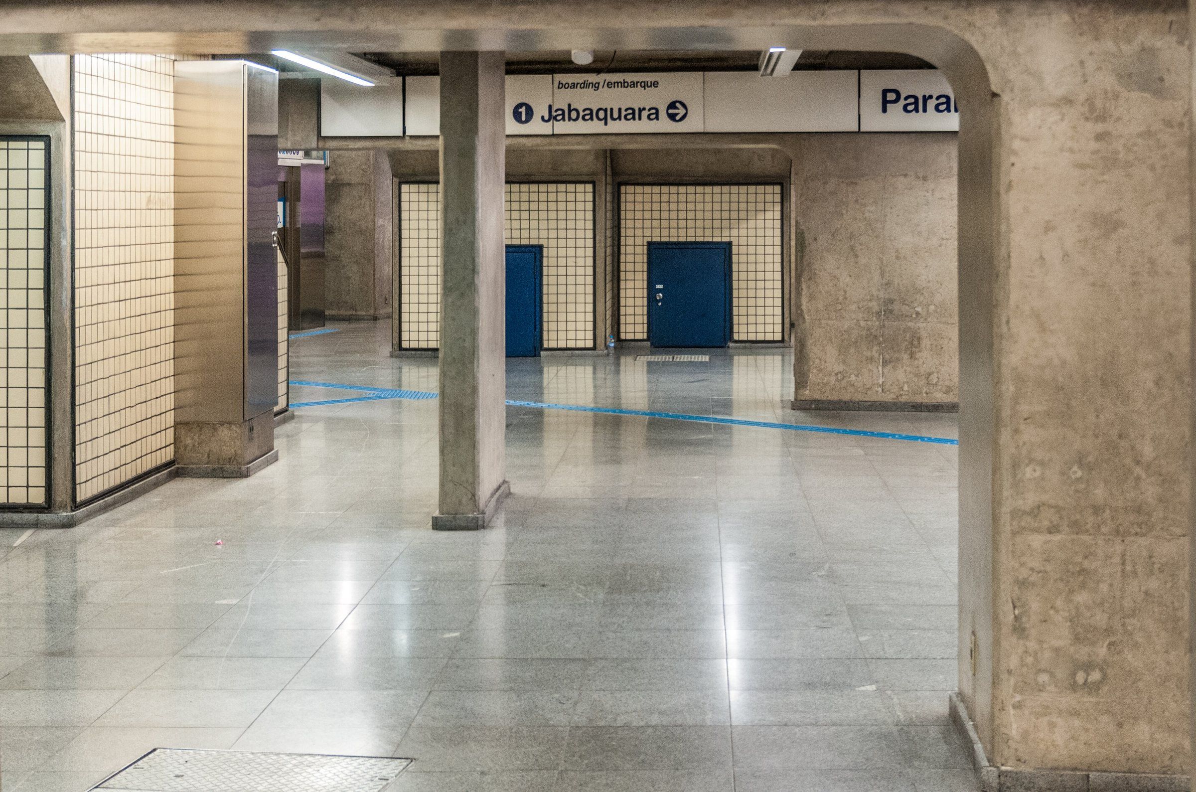 Mezanino vazio de uma estação de metrô: piso de granito polido, uma coluna de concreto isolada, paredes de azulejo atrás de grades, duas portas azuis fechadas e uma linha azul no chão que vira e some.