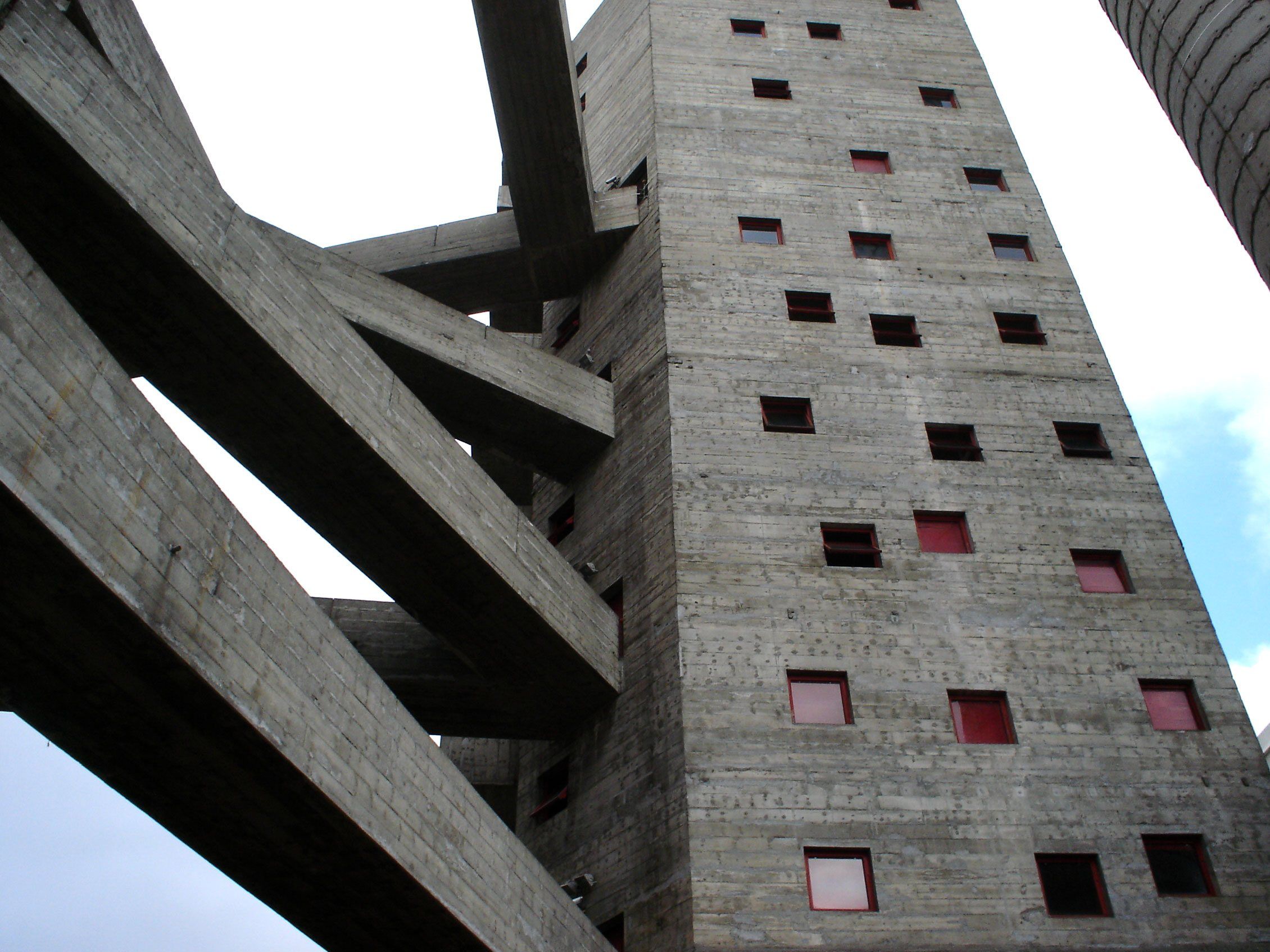 Vista de baixo de uma torre de concreto com pequenas janelas de moldura vermelha, cruzada por passarelas diagonais de concreto.